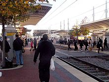 Numerous passengers walking and waiting along the tracks at the side platforms of the station with a train and arena at rear.