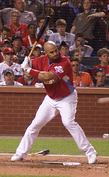 A right-handed batter is at the plate, looking toward the pitcher's mound. Wearing a red uniform and white pants, there is a crowd behind him with jerseys of various colors.