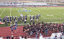 A high school football field with a large "DV" emblem in the center of the turf. Dougherty Valley football players in blue and silver uniforms are lined up on the side of the field. On the track surrounding the field, there are cheerleaders in light blue, white, and navy blue uniforms with blue pom-poms to the left. To the right of the track there are members of the pep band with their instruments in black shirts, surrounding music director Teri Musiel.