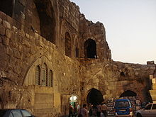 Cars and people in front of a stone wall with window and door openings and the remains of vaults