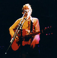 A male vocalist (wearing a cream shirt) singing into a microphone on stage while playing guitar