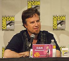 A Caucasian man in his forties, seated at a conference, with a microphone in front of him. He has a pleasant square face, deep-set eyes, dark hair and a brown beard with clean-shaved cheeks and upper-lip. He is casually dressed, relaxed and smiling. Square signs are posted on the wall behind him, bearing the name COMIC-CON in big bright yellow letters around a drawn eye and eyebrow.