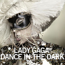 Face of a blond woman, whose platinum blond hair is shaped in a bob cut. She wears a shiny dark colored dress and has a black mask with strings around it on her eyes. The woman tilts her head to the right. Beneath her chin, the words "Lady Gaga" and "Dance in the Dark" are written in white bold font.