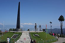 An obelisk on an elevated circular platform, with a few people standing around. Mountain peaks are visible in the background.