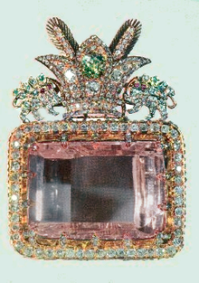 A large rectangular pink multifaceted gemstone, set in a decorative surround. The decoration includes a row of small clear faceted gemstones around the main gem's perimeter, and clusters of gems forming a crest on one side. The crest comprises a three-pointed crown faced by two unidentifiable animals.