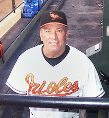 A man wearing a white baseball jersey with "Orioles" written across the chest in orange script and a black baseball cap with an orange oriole on the front