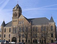 A large three-story stone building built in 1895. Three stories of windows line the front of the building with the two front corners containing cone-shaped roofs that stick out from the main roof. Above the entrance is a large clock tower that is taller than the rest of the building.