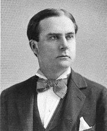 A man with black hair wearing a black jacket and vest, patterned bowtie and white shirt and pocket square