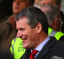 A side-on photograph of a man with greying hair. He is wearing a light blue shirt with a red tie and a dark jacket.