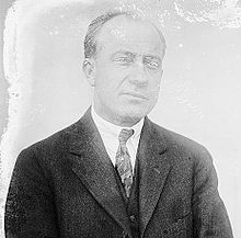A man with graying, receding hair wearing a dark jacket, patterned tie and white shirt