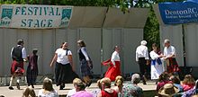 Four couples dressed in Celtic costumes dance on stage on a sunny day with audience in seats looking on.