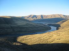 A relatively small river winds through a series of sparsely vegetated brown hills under a nearly cloudless blue sky. The river and the low hills are in shade, while the hilltops and a large hillside in the foreground are sunlit.