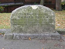 A stone gravestone with a curved top, with several lines of inscription.  Trees, grass, and a wall are visible in the distance.