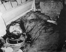 Middle-aged black-haired man lies face half-down on the floor, covered on his face and dark suit and trousers with blood. His hands are behind his back.