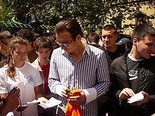 A dark-haired man in sunglasses and a wide-collared open shirt holds a pen in his hand as he looks down at a piece of paper held out to him by one of a crowd of people which surrounds him.