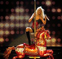 Blonde woman wearing a red and black teddy and black boots, holding a microphone, and stepping onto carousel horse.