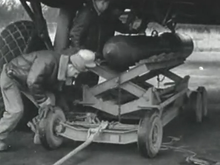 Photograph of three men using lifting equipment to jack a heavy bomb up to the underside of a large parked aircraft