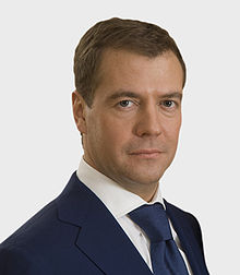 A portrait shot of a serious looking middle-aged male looking straight ahead. He has short brown hair, and is wearing a blue blazer with a blue tie over a white collared shirt.