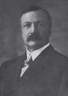 A man with dark, receding hair and a thick mustache, wearing a white shirt, colored tie, and dark jacket