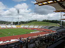 Interior of a sports stadium. There is a running track surrounding a central grassed area. In the distance there are the outer stands. In the foreground is the main stand, with plenty of people seated