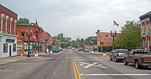 A wide street with a double yellow line and buildings on either side, more on the left than the right. American flags are on the lampposts, and signs both on the road and next to it indicate the railroad crossing in the distance.