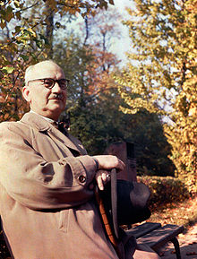 An elderly man, wearing a grey coat and holding a black hat sits in a garden in autumn.