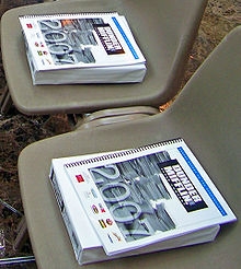 A book titled "Dunder Mifflin 2007 Annual Report" on top of a ream of paper in two empty plastic chairs