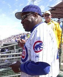An older African American man wearing a white, pinstriped baseball uniform and sunglasses; he is autographing a baseball at a baseball game
