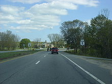 Ground-level of two lanes of a divided highway with a small grassy median; the highway intersects with another road at a traffic signal that is visible in the distance.
