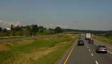 A four lane divided highway among short hills travels into the background and curves to the right. The two divided halves are separated by a depressed swampy median.