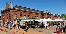 A wide, one story red brick building with a sloping black roof behind numerous white-topped tents selling assorted wares.
