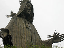 A metal statute of a woman wearing a hooded cloak with a kindly expression whose shoulder and outstretched hand are perched on by stylized birds