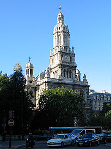 A 19th-century church in the French style, in light coloured stone, with a central tower with rounded top and smaller towers set back to left and right. The sky is blue. There are trees in leaf, and some cars parked in front of the church.