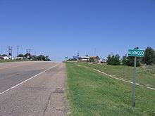 A small green sign reading "ELMWOOD". In the background is a highway intersection with beacons. Nearby are two abandoned gas stations.