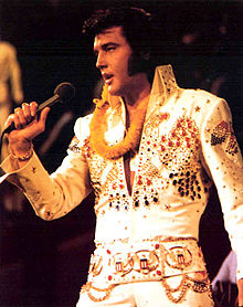 Presley, mutton-chopped and fuller-faced, sings into a handheld microphone. A golden lei is draped around his neck, and he wears a high-collared white jumpsuit resplendent with red, blue, and gold bangles.