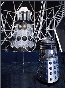 Full length image of the Dalek Emperor, standing upon a dark blue plinth, with a light grey geodesic framework in the background and a Mark 3 Dalek in the foreground.
