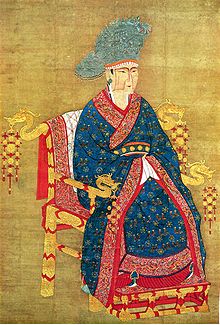 A painting of a woman in a blue dress with intricate gold and red decorations and a large blue hat. Her face has a simple angular design painted onto it in darker brown tones. She is sitting in a golden throne with dragon heads protruding from the ends of the armrests and from the sides of the top, back edge of the throne.