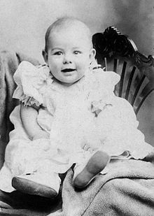  an infant dressed in light colored clothing propped on a chair faces the camera