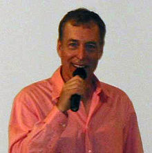 A man with short dark hair in a pink shirt, speaking into a microphone in his right hand.
