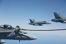 Three grey jet fighters in formation. The aircraft closest to the camera has a hose attached to it.