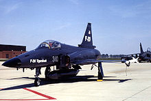 Dark-blue jet aircraft taxiing on ramp, carrying an external fuel tank under belly.