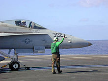 Side nose view of bluish gray jet aircraft on aircraft carrier out at sea on clear day. Green-topped personnel wearing earmuffs standing close by, preparing the aircraft for launch towards right