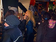 Blond woman having make-up applied on a movie set, surrounded by busy people.