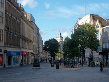 Fargate shopping precinct, Sheffield. Once a busy road, it has been pedestrianised for several decades and is Sheffield's main City Centre shopping area, home to many well known companies. The image shows classical architecture on both sides with one plan spaces in the centre, dotted with trees and the buildings on the High Street are visible beyond the trees.