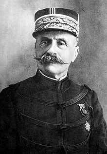 An elderly gentleman with a handlebar-style moustache. He is wearing a peaked cap highly decorated with light-coloured braiding. He wears a dark, high-collared jacket, with regularly spaced horizontal bands of braiding. On his left breast, he wears a medal on a ribbon above a star.