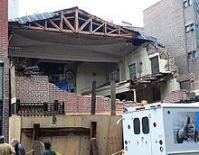 A building is surrounded on the first floor by plywood hoarding. The second and third floors are partially open to the street, and the interior can be seen. Part of the roof has also been torn away, and the joists and trusses are exposed.