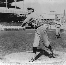 A man wearing a baseball uniform stands truned to his right having just pitched a baseball from the pitcher's mound.
