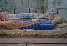 Two above ground tombs with reclining statues on top. The near tomb has a male crowned figure holding a sceptre, with his robes painted blue and red. The far tomb has a female crowned figure holding an open book with her robes painted blue.