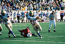 Several men dressed in football uniforms dart down a field. Another football-suited man is collapsed on the ground, while a man is scene in the background wearing a black and white striped shirt holding a whistle. Behind the scene are hundreds of people cheering in stands.