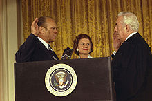 A man in a suit, his right hand in the air, stands next to his wife and speaks to another man in the robes of a judge. The group stands in front of a curtain, behind a podium bearing the seal of the President of the United States.